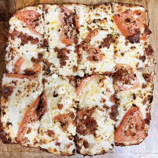 Square-cut Hindsight Pizza with white cheese, crumbled sausage, and tomato slices on a wooden board.