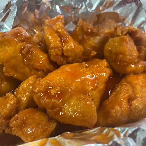 Close-up of golden-brown boneless wings coated in a glistening orange sauce, served in a foil container.