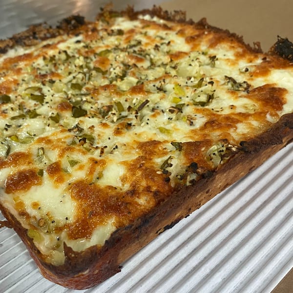A rectangular Hindsight Pizza with melted cheese, green peppers, and black seasoning on a white surface.