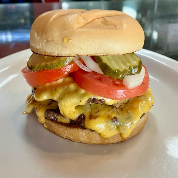 A double cheeseburger with melted cheese, tomatoes, pickles, and onions on a white plate.